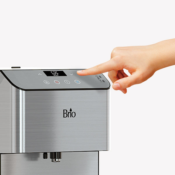 A hand presses a button on the control panel of a Brio water dispenser with a digital display and stainless steel finish.